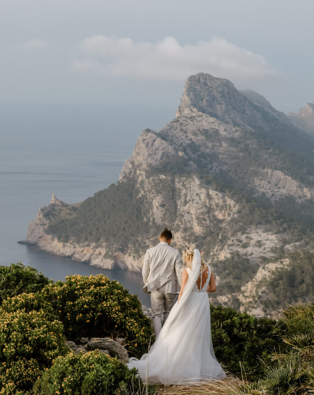 Bride and Groom in wedding attire from the back standing on cliffs of Cap de Formentor Mallorca with cliffs and Mediteranean Sea in the background