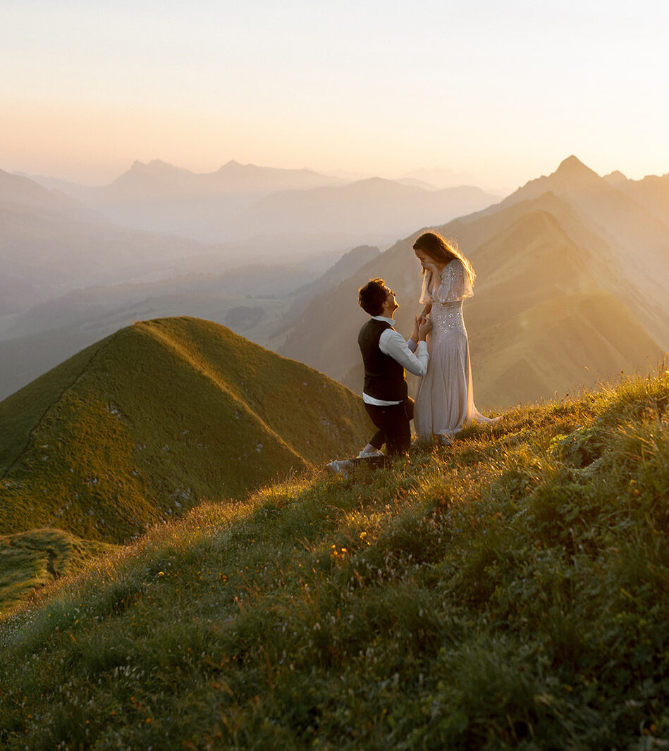 Couple on a mountain ridge at sunrise, man on one knee proposing to surprised women in a dress