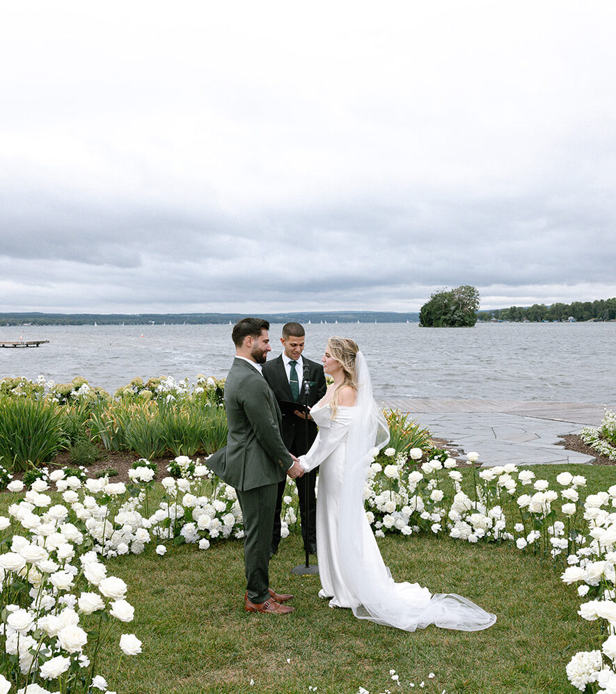 Couple exchanging vows with an officiant during their outdoor wedding ceremony on a meadow decorated with white florals with Canandaigua lake in the background