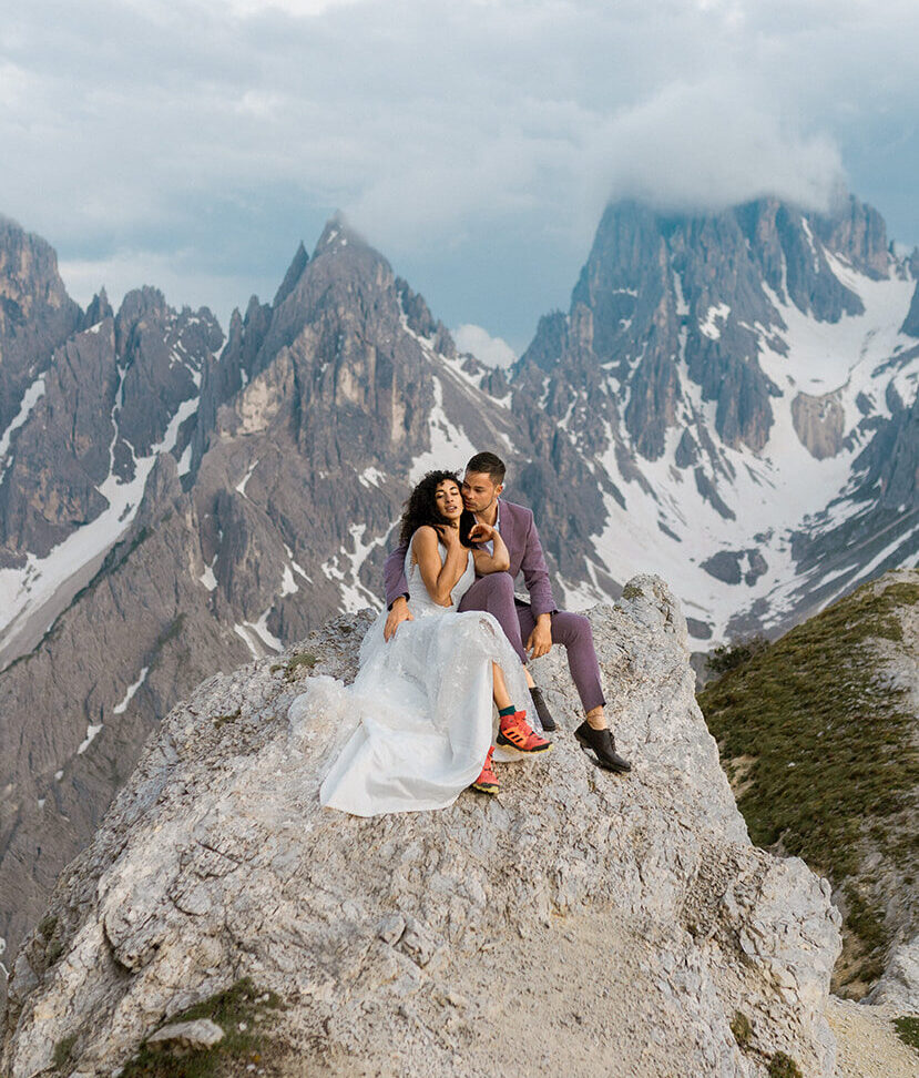 A couple in wedding attire during their elopement in the Dolomites sitting on a big rock with the Cardini di Misurina towering in their background